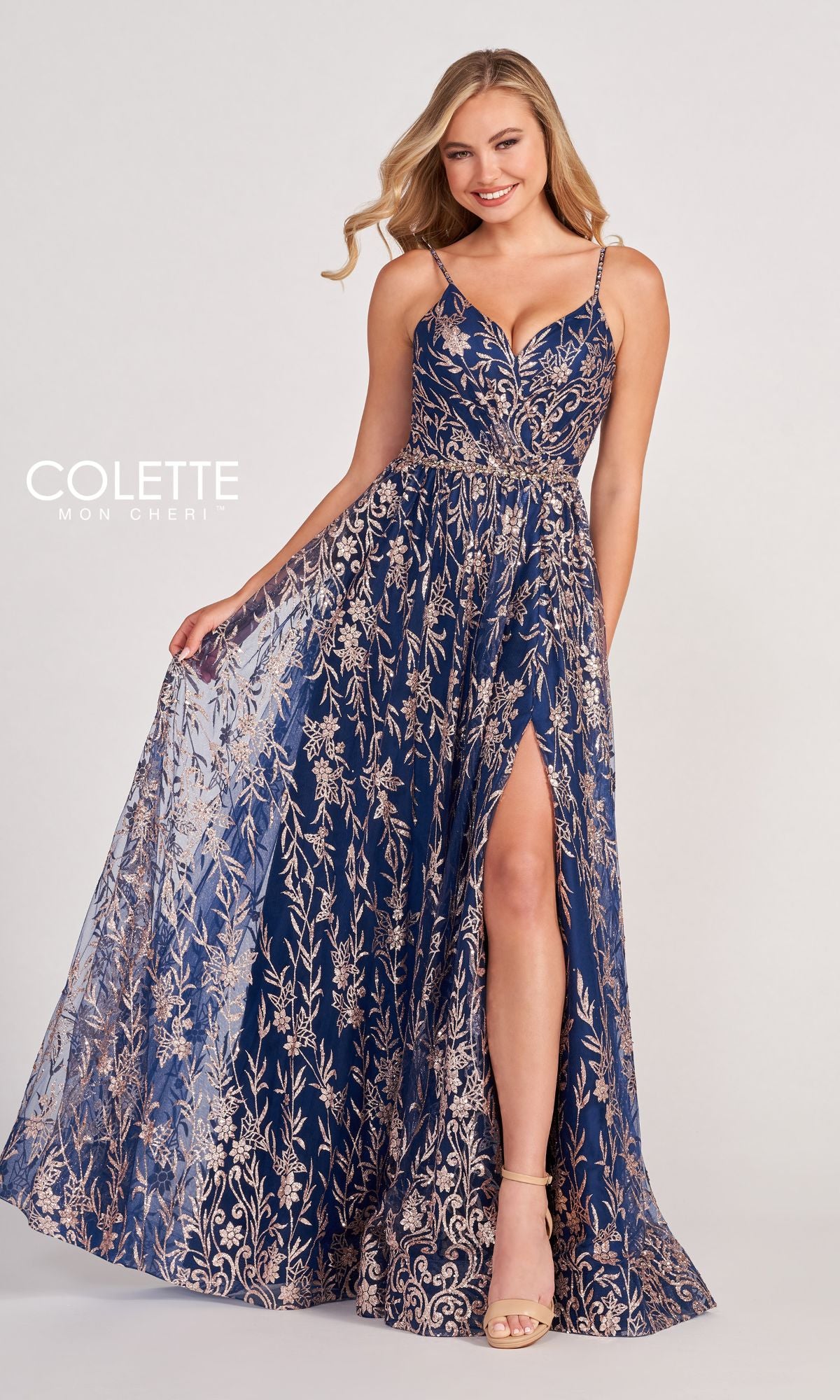 A woman poses and smiles in the Colette CL2022 Strappy A-Line Glitter Prom Dress, featuring a navy blue and gold floral pattern, backless design, and high slit against a plain background.